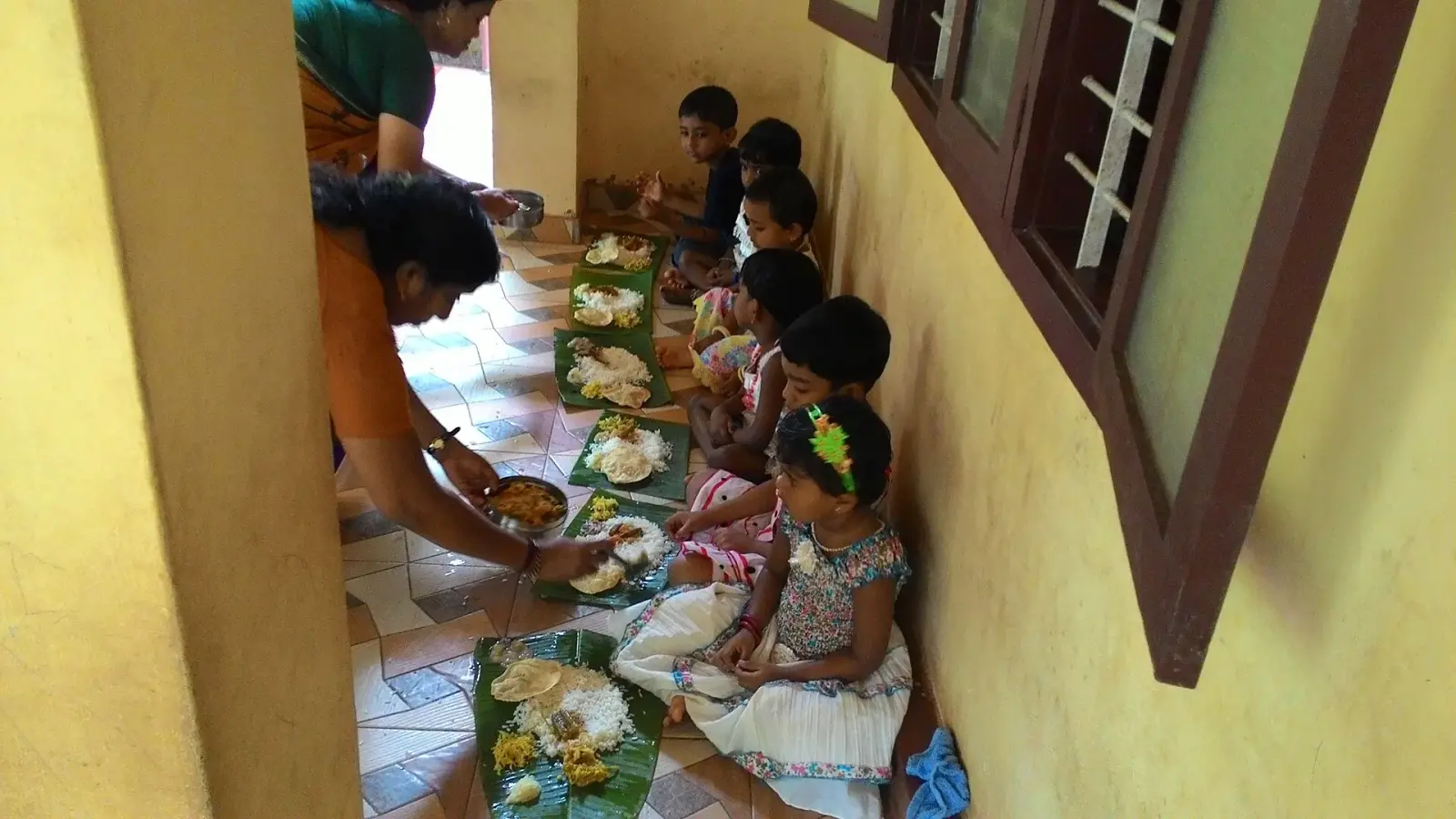 A volunteer serving a meal on banana leaves to children seated in a row